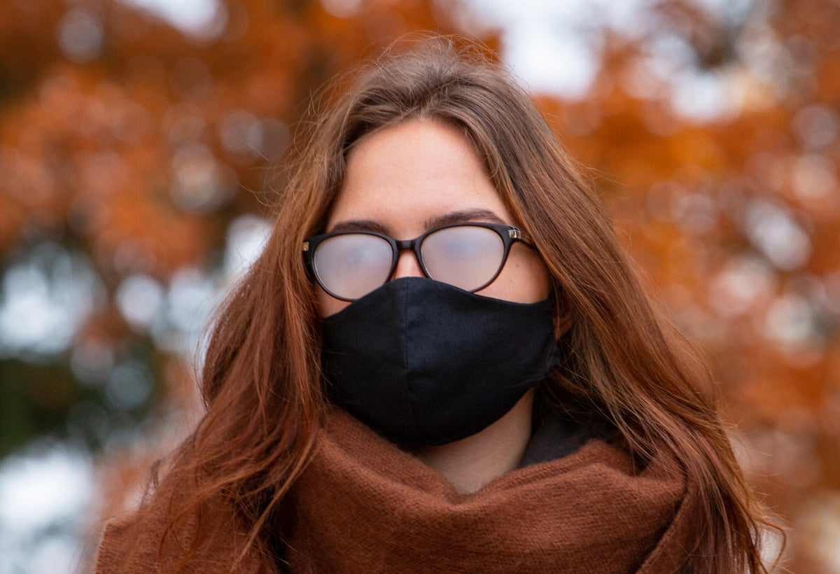 A woman outdoors in cool weather wearing a black fabric face mask, her spectacle lenses completely fogged over